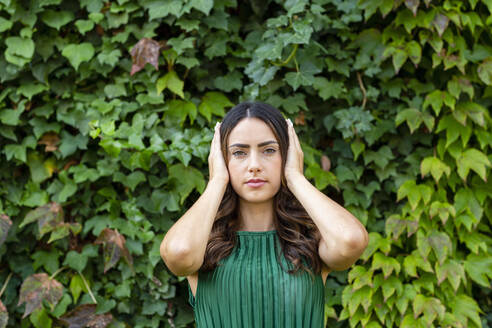 Schöne Frau hält sich die Ohren mit den Händen vor grünen Efeupflanzen zu - EIF02216