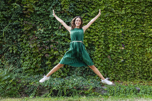 Playful woman with arms outstretched jumping in front of ivy plants at park - EIF02220