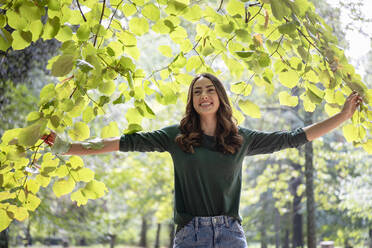 Happy young woman with arms outstretched holding branches - EIF02226