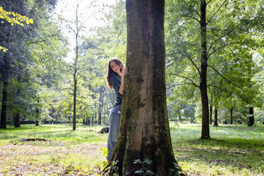 Smiling woman standing behind tree trunk in park - EIF02238