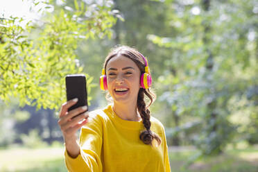 Happy woman with headphones taking selfie through smart phone at park - EIF02256