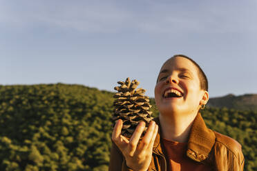 Happy woman holding pine cone during sunny day - MGRF00516
