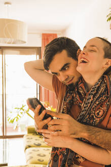 Mann benutzt Mobiltelefon, während er eine Frau zu Hause umarmt - MGRF00524