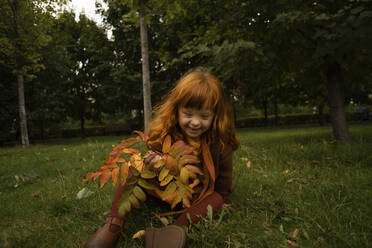 Lächelndes rothaariges Mädchen mit Blättern auf Gras im Park sitzend - SSGF00046