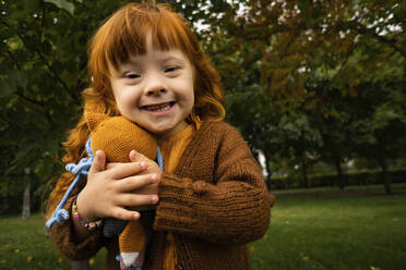 Lächelndes Mädchen mit Down-Syndrom umarmt Spielzeug im Park - SSGF00049