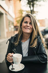 Lächelnde reife Frau hält Tasse, während sie in einem Straßencafé sitzt - GRCF01014