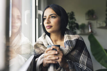 Junge Frau mit Kaffeetasse schaut durch das Fenster zu Hause - JCCMF04199