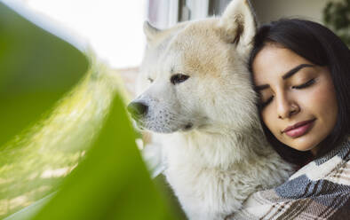 Beautiful woman with eyes closed embracing Akita dog by window - JCCMF04204