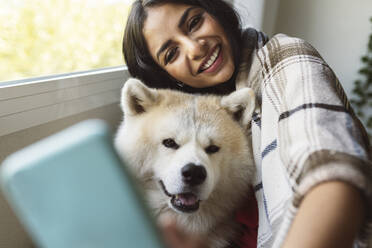 Smiling woman with Akita dog taking selfie through mobile phone at home - JCCMF04206