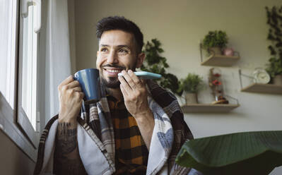 Lächelnder Mann mit Kaffeebecher in der Hand, der am Fenster mit seinem Handy telefoniert - JCCMF04211