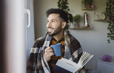 Smiling man with book having coffee during winter at home - JCCMF04212