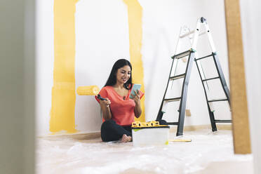 Smiling young woman with paint roller using smart phone in living room - JCCMF04216