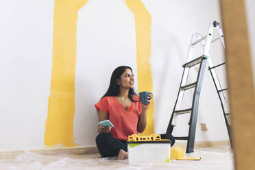 Young woman having coffee while holding mobile phone in living room - JCCMF04217