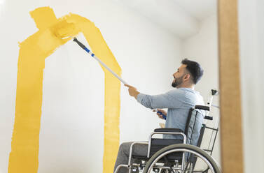 Mann sitzt im Rollstuhl und streicht die Wand im Wohnzimmer - JCCMF04219