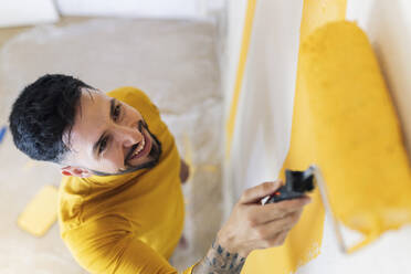 Smiling man with paint roller painting wall in living room - JCCMF04225