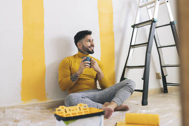 Lächelnder Mann im mittleren Erwachsenenalter, der zu Hause auf einer Leiter Kaffee trinkt - JCCMF04226