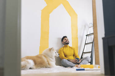 Smiling man sitting cross-legged by dog at home - JCCMF04230
