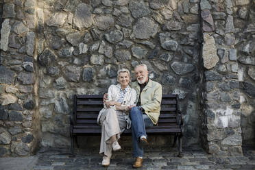Lächelndes Paar sitzt mit gekreuzten Beinen vor einer Steinmauer - LLUF00188