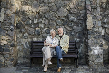 Smiling couple sitting with legs crossed at knee in front of stone wall - LLUF00188