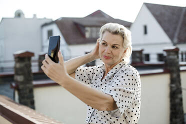 Senior woman taking selfie through smart phone on rooftop - LLUF00200