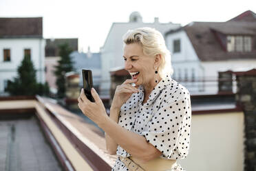 Glückliche Frau, die an einem Videoanruf über ein Smartphone auf einer Gebäudeterrasse teilnimmt - LLUF00201