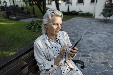 Ältere Frau benutzt ein Mobiltelefon, während sie auf einer Bank sitzt - LLUF00207