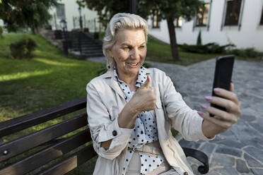 Lächelnde Frau, die während eines Videoanrufs über ein Mobiltelefon auf einer Bank die Daumen nach oben streckt - LLUF00208