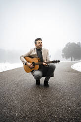 Lächelnder Mann spielt Gitarre auf der Straße im Winter - RCPF01339