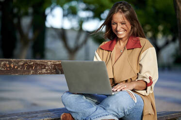Glückliche Frau, die einen Laptop benutzt und im Schneidersitz auf einer Bank sitzt - KIJF04176