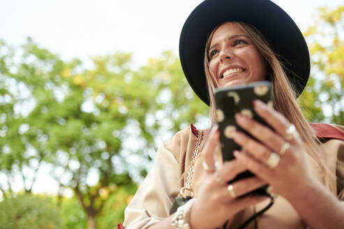 Lächelnde Frau mit Hut, die wegschaut und ein Smartphone hält - KIJF04184