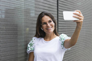 Lächelnde weibliche Fachkraft, die ein Selfie durch ein an die Wand gelehntes Mobiltelefon macht - PNAF02500