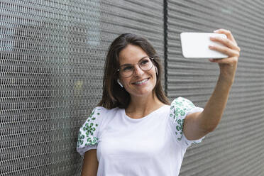 Smiling female professional taking selfie through mobile phone leaning on wall - PNAF02500