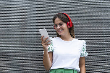 Glückliche junge Geschäftsfrau, die ein Smartphone benutzt, während sie vor einer Wand Musik hört - PNAF02505
