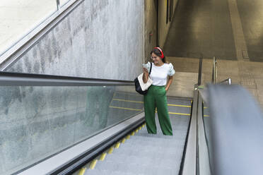 Junge Berufstätige, die auf einer Rolltreppe stehen und mit ihrem Smartphone Musik hören - PNAF02508
