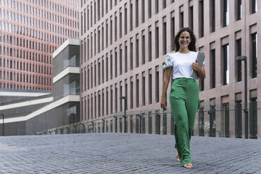 Smiling young businesswoman walking with laptop on footpath - PNAF02519