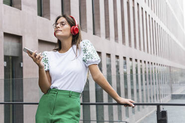 Junge Geschäftsfrau mit geschlossenen Augen, die ihr Smartphone hält, während sie an einem Glasgeländer Musik hört - PNAF02530