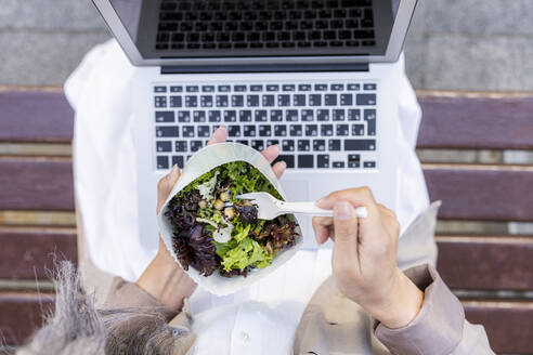 Frau mit Laptop isst Salat und sitzt auf einer Bank - OIPF01310