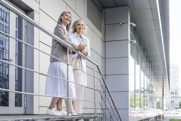 Geschäftsfrauen schauen weg, während sie an einem Geländer vor einem Bürogebäude stehen - OIPF01323