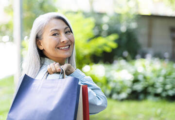 Happy mature woman looking over shoulder while carrying shopping bags - OIPF01340