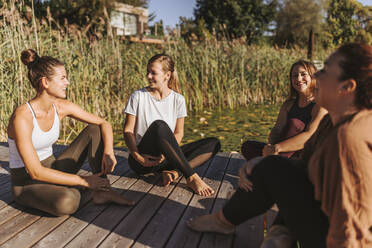 Female friends talking to each other while sitting on jetty by lake - DAWF02036