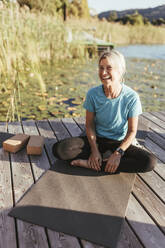 Glückliche Frau, die wegschaut, während sie beim Yoga-Kurs auf einer Übungsmatte sitzt - DAWF02053