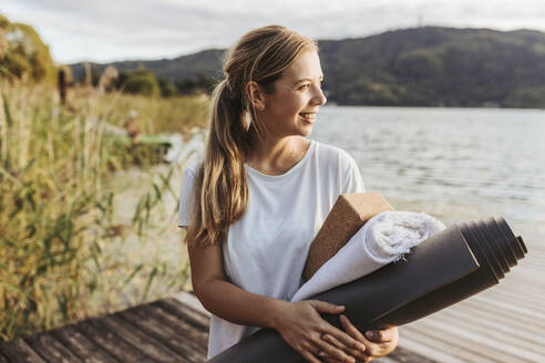 Lächelnde Frau schaut weg und hält eine Gymnastikmatte am See - DAWF02058