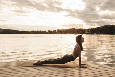 Woman practicing yoga on jetty by lake during sunset - DAWF02059