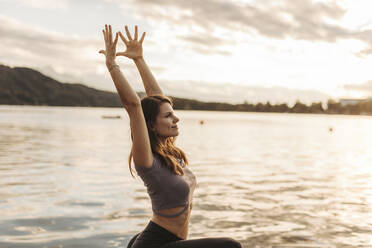 Woman with arms raised doing yoga by lake during sunset - DAWF02060