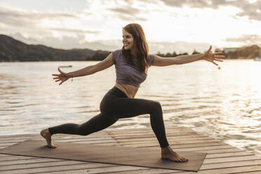 Smiling woman with arms outstretched doing yoga on jetty during sunset - DAWF02061