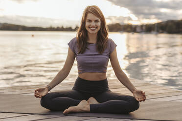 Smiling woman practicing lotus position while sitting on exercise mat by lake during sunset - DAWF02063