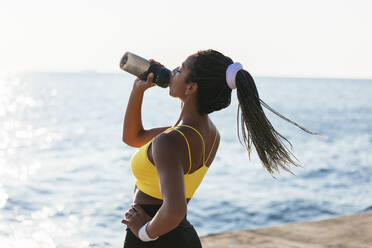 Young sportswoman drinking water while standing near sea - OYF00596