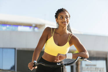 Lächelnde junge Sportlerin mit Fahrrad träumt an einem sonnigen Tag - OYF00604