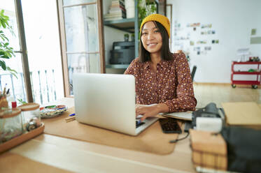 Berufstätige Frau mit Strickmütze sitzt mit Laptop am Arbeitsplatz - KIJF04219