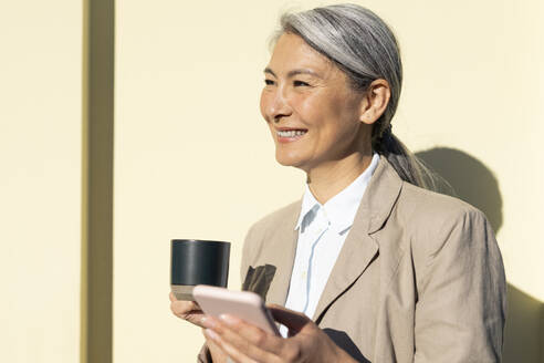 Glückliche reife Frau mit Kaffeetasse und Smartphone vor einer Wand stehend - OIPF01349